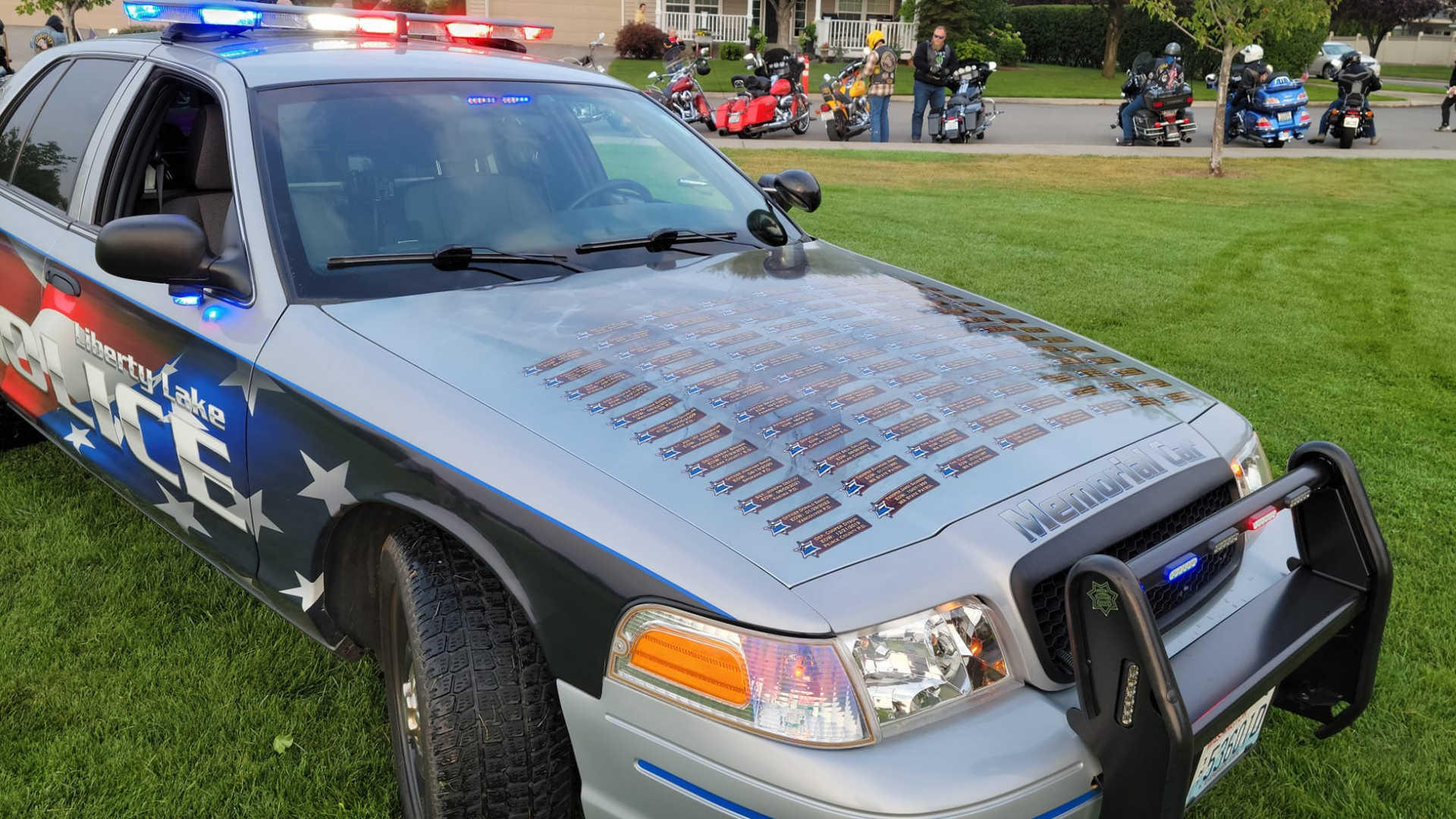 Image of a police car from Liberty Lake Police with emergency lights on, parked on grass, displaying a hood covered with memorial plaques at dusk.