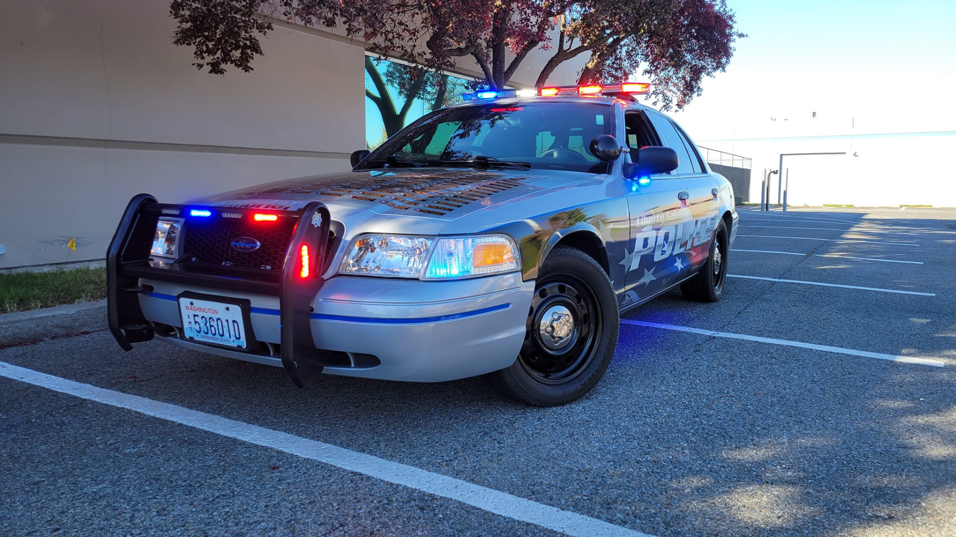 Close-up view of a Liberty Lake Police patrol car with flashing red and blue lights, showing a hood filled with police memorial plaques in daylight.