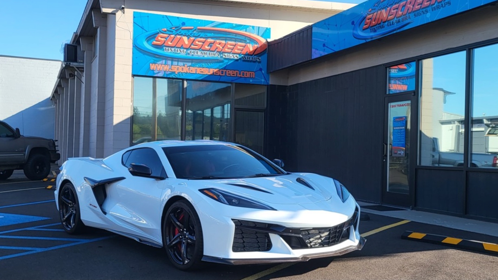 A sleek white Chevrolet Corvette parked in front of Spokane Sunscreen signage, with blue sky reflecting on the car's shiny surface.