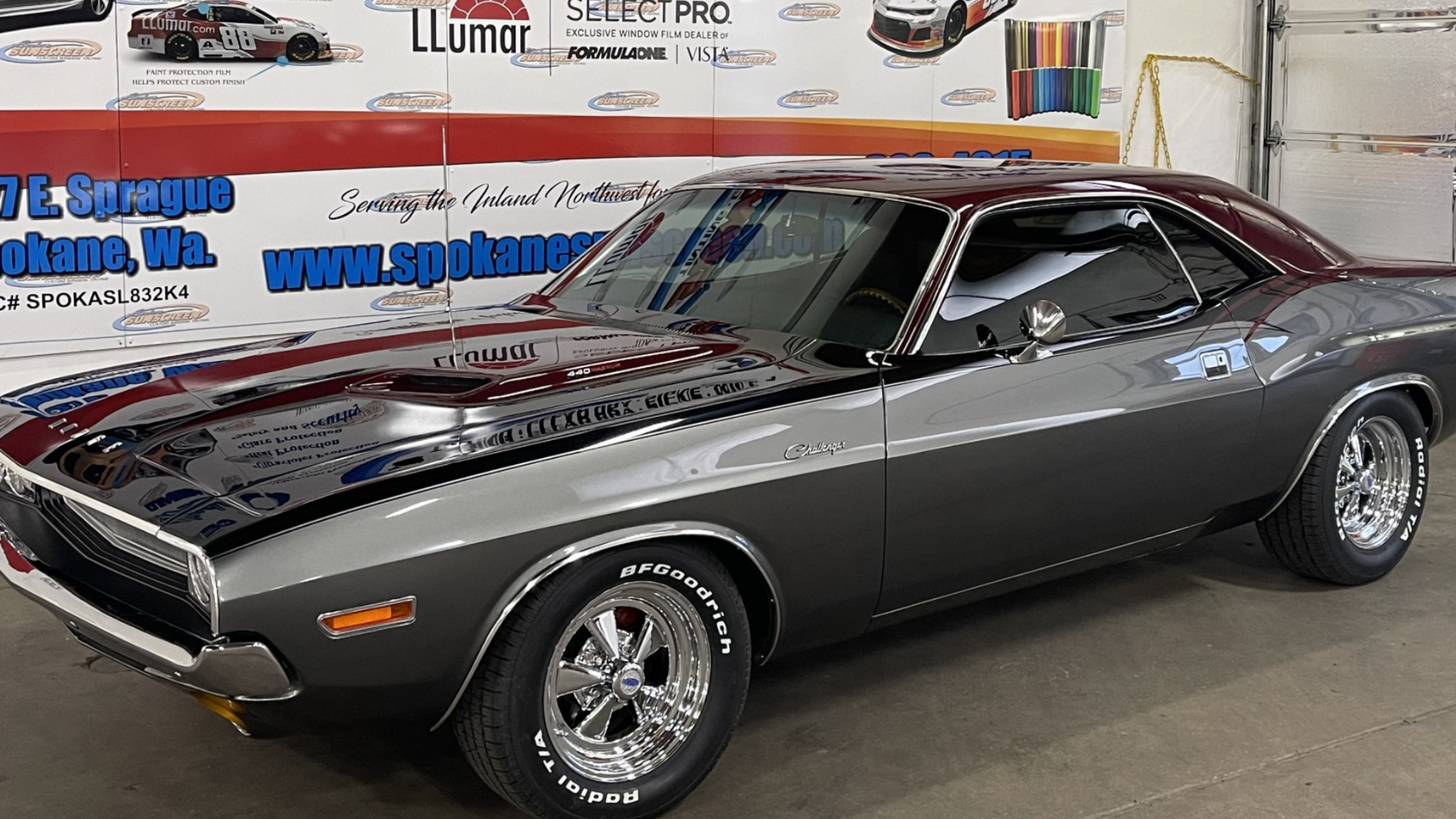 Classic silver Dodge Challenger with black stripes on display inside a showroom, featuring chrome wheels and the Challenger emblem on the side.