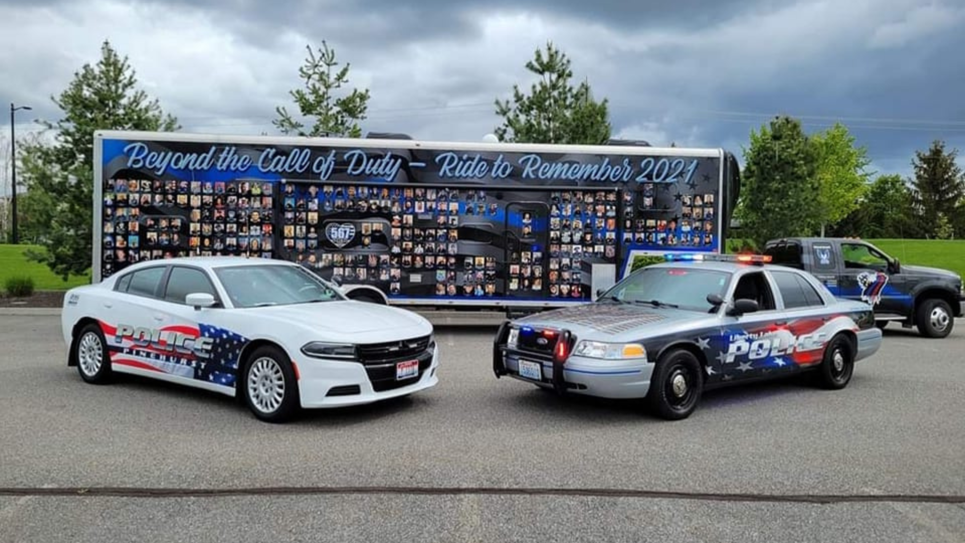 A wide shot featuring two police vehicles, one from Liberty Lake Police, in front of a trailer displaying photos and names of officers lost in the line of duty, titled "Beyond the Call of Duty – Ride to Remember 2021.