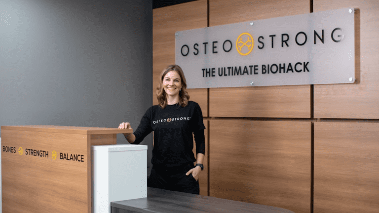 A smiling woman stands behind a reception desk at OsteoStrong, with a sign behind her that reads "OsteoStrong: The Ultimate Biohack." The desk has the words "Bones," "Strength," and "Balance" on the front.