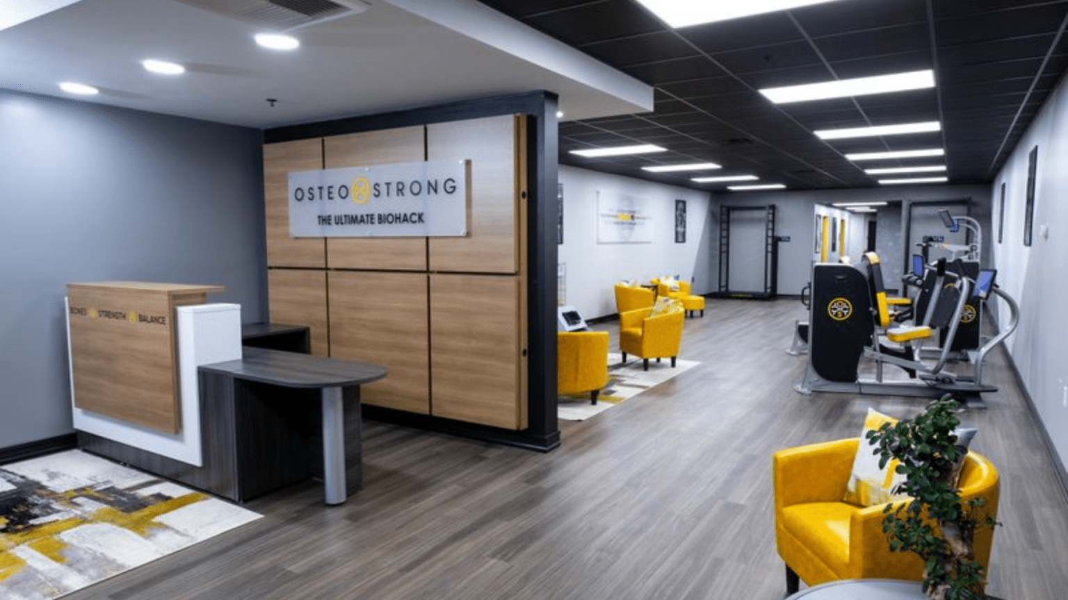 The reception area of OsteoStrong, featuring a modern, clean design with wood accents, a reception desk, and seating areas with yellow chairs. Exercise equipment is visible in the background.