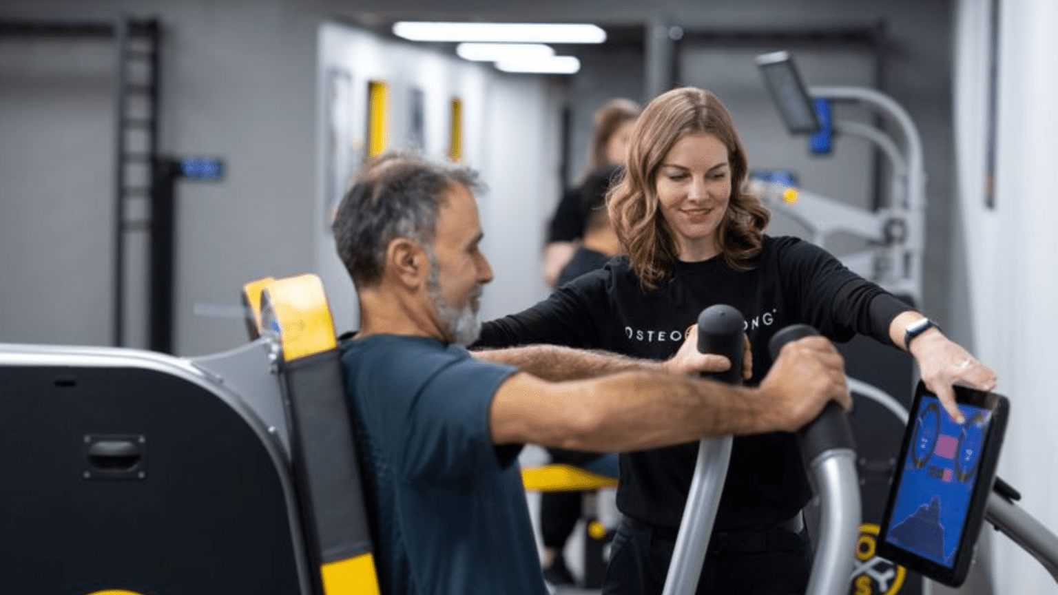 A woman in an OsteoStrong shirt assists an older man using exercise equipment, monitoring his progress on a screen.