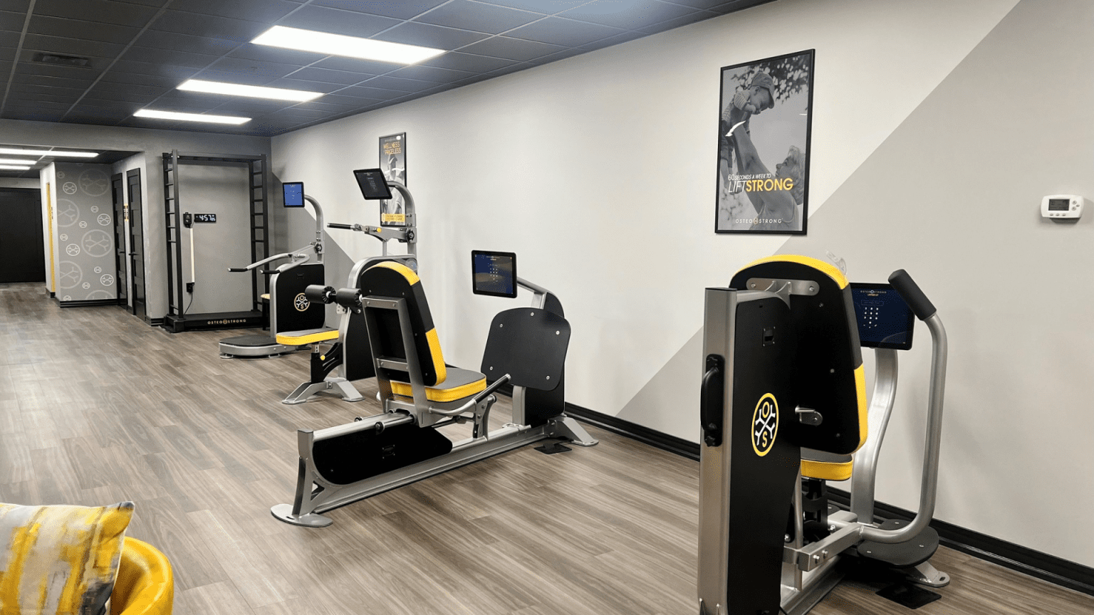 A spacious exercise area at OsteoStrong with various pieces of exercise equipment. The walls are decorated with motivational posters, and yellow chairs are arranged in a seating area.