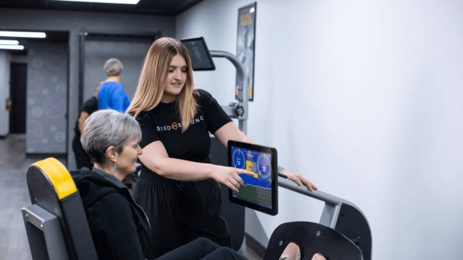 A young woman in an OsteoStrong shirt explains the use of exercise equipment to an older woman, pointing to a screen displaying data.