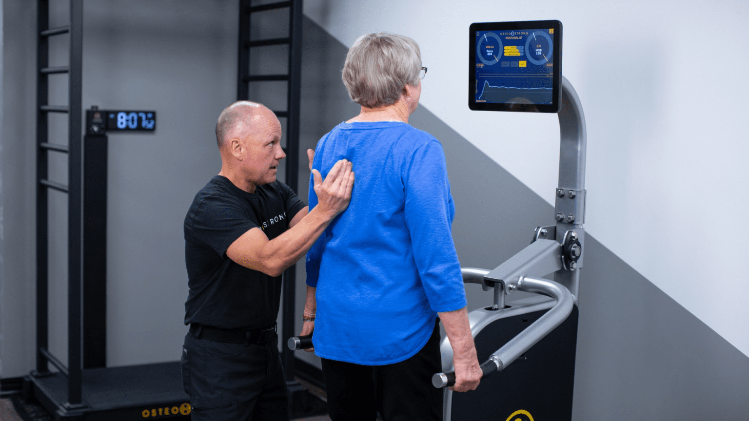 A man in an OsteoStrong shirt helps an older woman use an exercise machine, demonstrating proper technique. The screen on the machine displays exercise metrics.