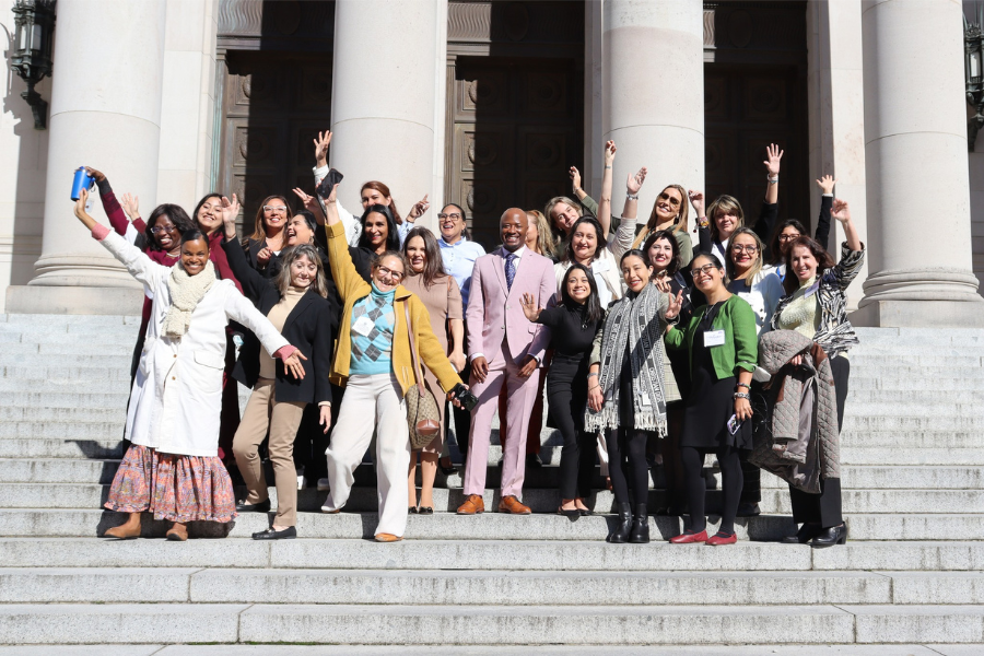 Group of diverse individuals standing on the steps of a building with large columns, smiling, and raising their hands in celebration. The group includes people of various ages and ethnicities.