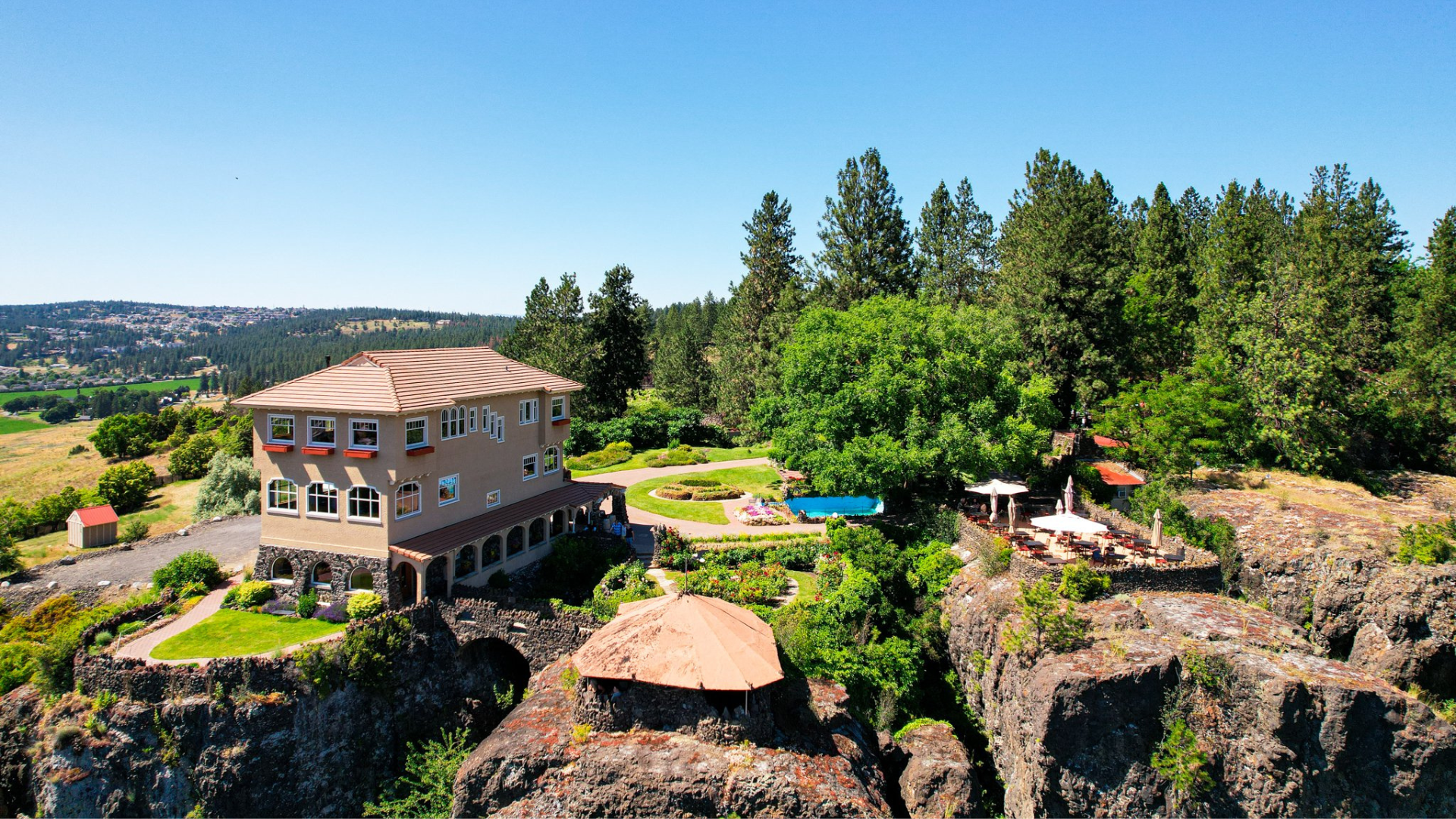 **ALT text:** Aerial view of a large, beige house with multiple windows and a tiled roof situated on a rocky cliff. The property features a well-manicured garden with circular pathways, a swimming pool, and a patio area with umbrellas and seating. Surrounding the house are lush green trees, and in the distance, rolling hills and a town can be seen under a clear blue sky.
