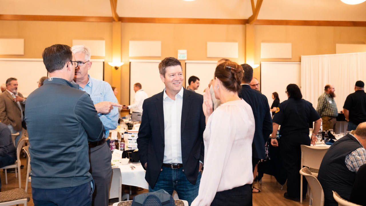 A group of professionals engage in conversation at a networking event. The room is filled with people in business attire, discussing and connecting with each other. The atmosphere appears friendly and collaborative, with a focus on building relationships. The event is held in a warmly lit room with tables set up for discussions.