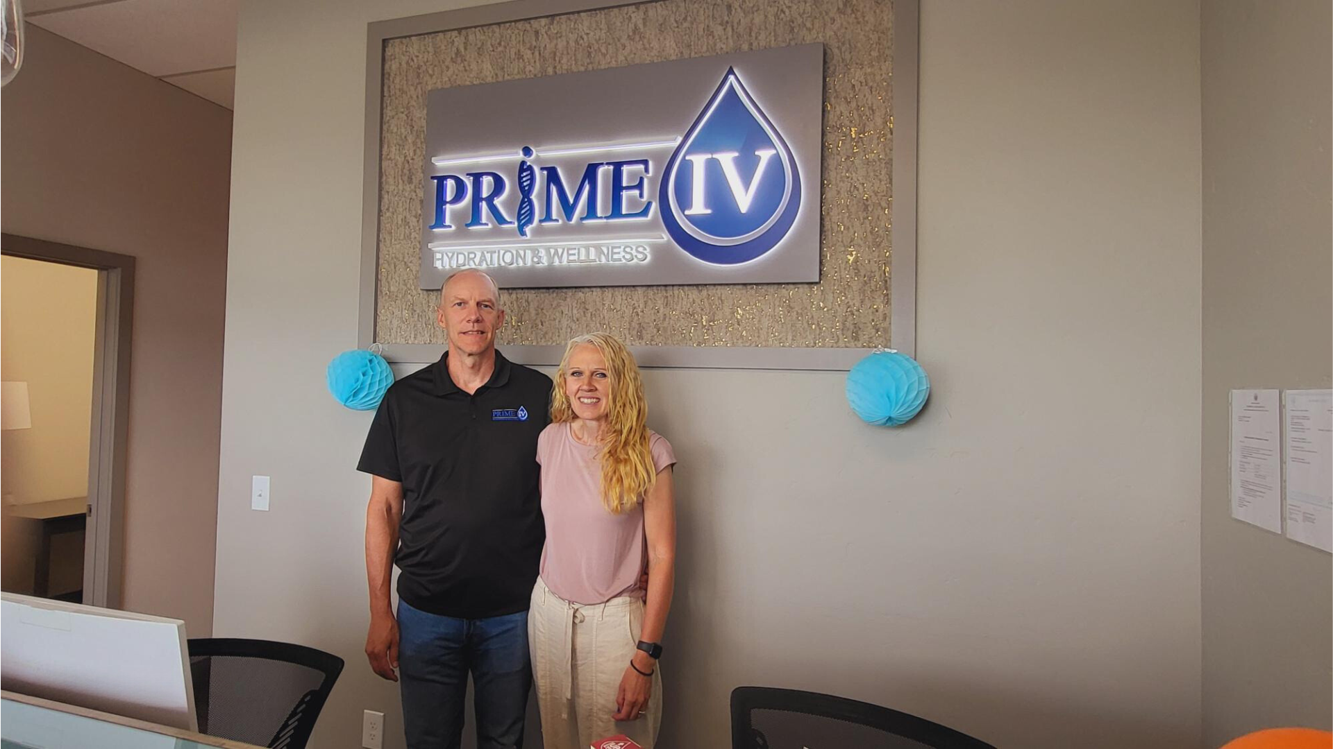 A man and a woman smiling while standing in front of a "Prime IV Hydration & Wellness" logo displayed on a wall inside a wellness clinic.