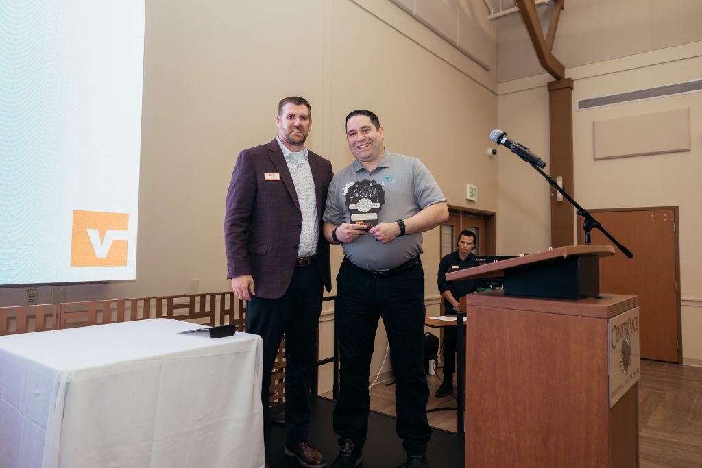 Award recipient holding manufacturing excellence trophy stands with presenter in burgundy blazer at podium event, with Chamber of Commerce logo displayed on screen in background.