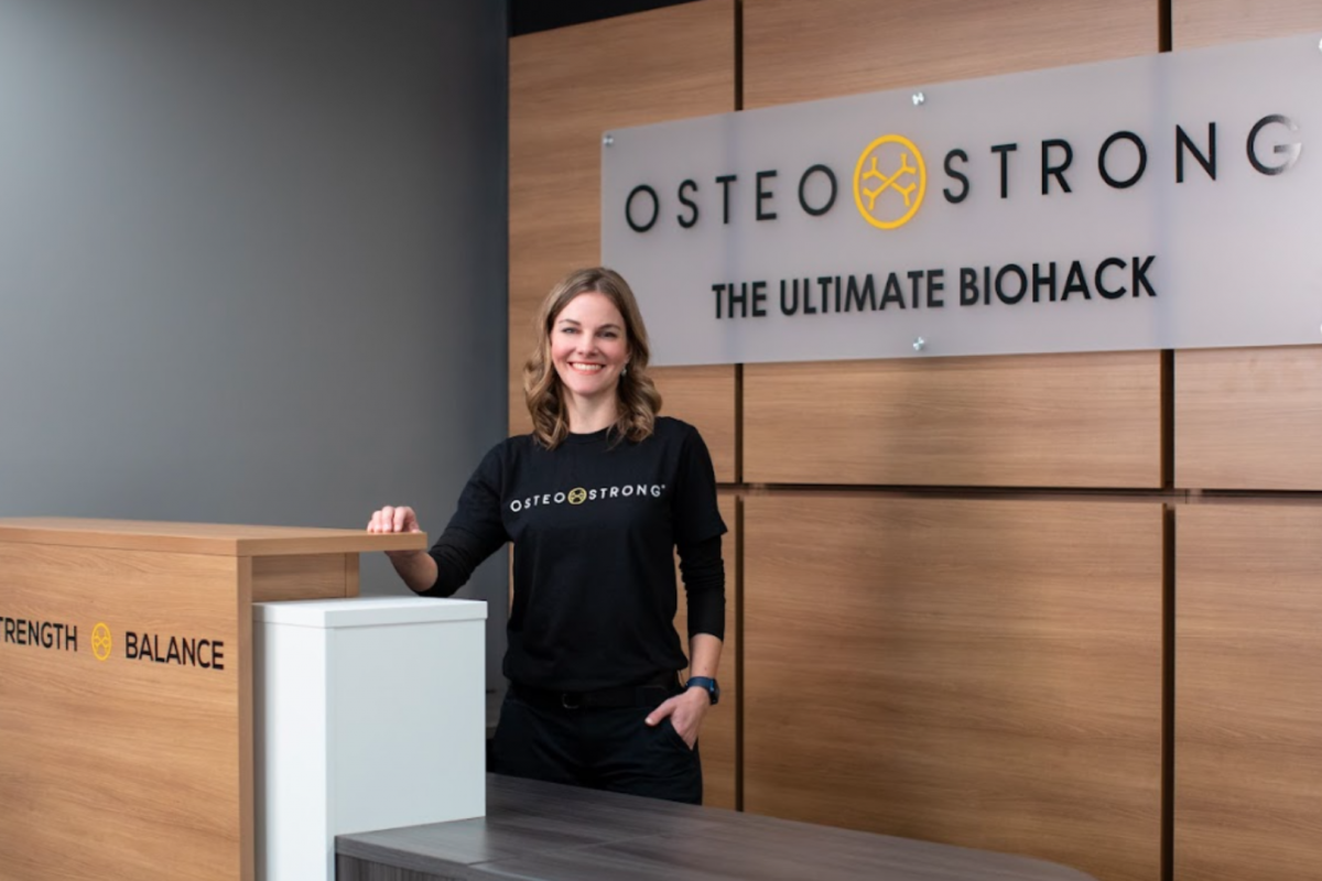 A smiling woman stands behind a reception desk at OsteoStrong, with a sign behind her that reads "OsteoStrong: The Ultimate Biohack." The desk has the words "Bones," "Strength," and "Balance" on the front.