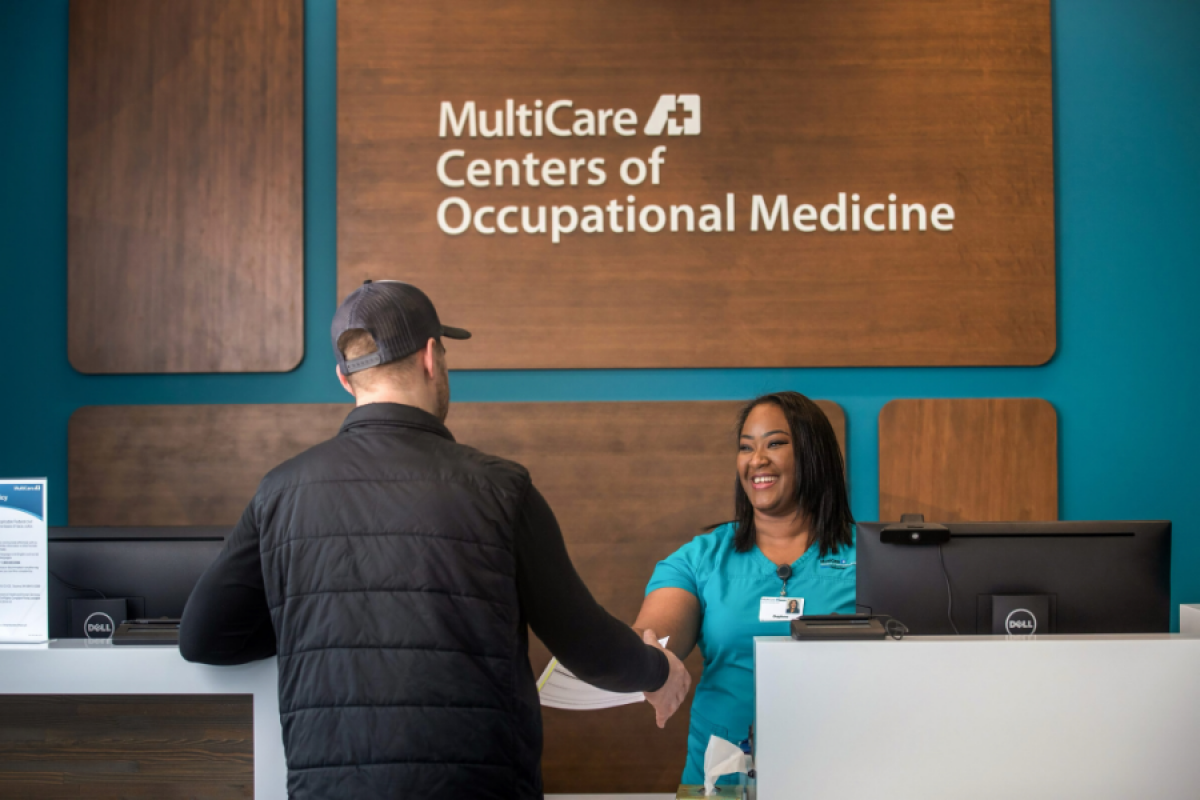 A friendly receptionist greets a patient at the front desk of MultiCare Centers of Occupational Medicine, highlighting professional healthcare services in a modern clinic setting.